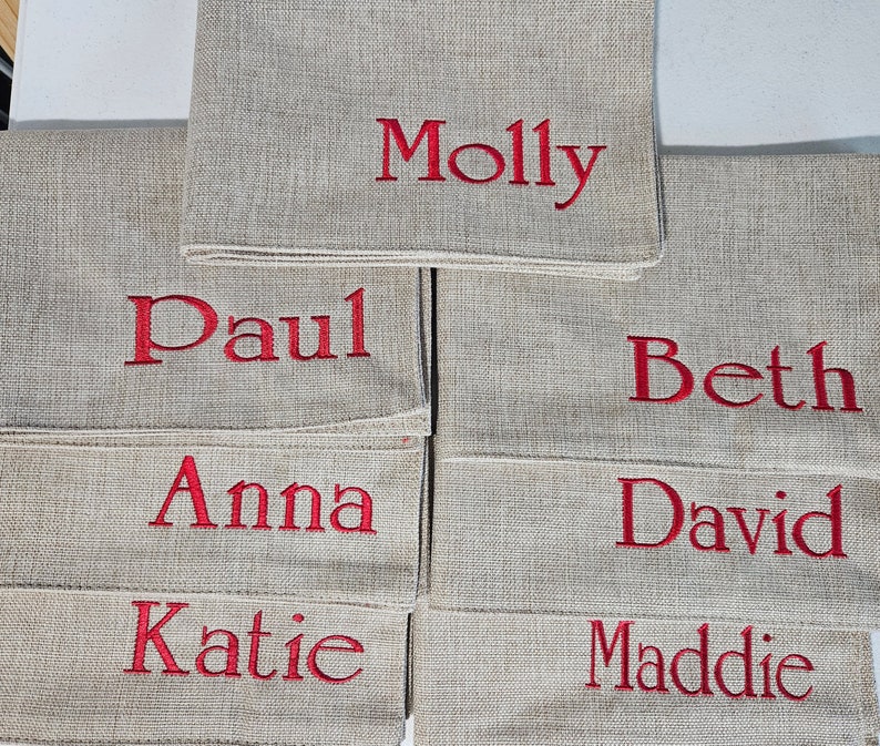Op de afbeelding: Een set van tien beige linnen placecards met rode geborduurde namen: Molly, Paul, Beth, Anna, David, Katie en Maddie.