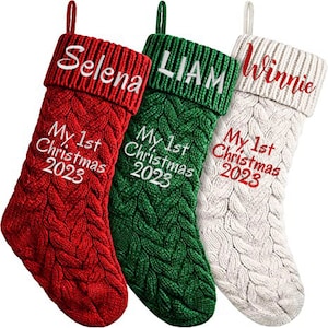 May include: Three red, green, and white knitted Christmas stockings with the names "Selena", "Liam", and "Winnie" embroidered on the top and the text "My 1st Christmas 2023" embroidered on each stocking.