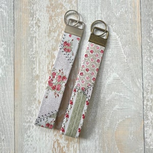 May include: Two fabric keychains with floral patterns. One keychain has a white background with pink and red roses and green leaves. The other keychain has a white background with pink and green floral patterns. Both keychains have silver key rings.