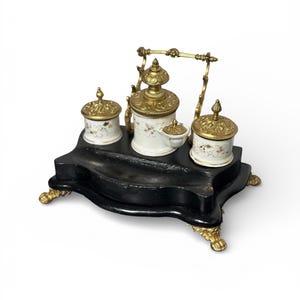 Peut inclure: Un ensemble d'encrier antique avec une base en bois noir et des accents dorés. L'ensemble comprend trois pots à encre en porcelaine blanche avec des couvercles dorés et des motifs floraux, et un pot à encre central avec un couvercle à charnière. La base a des pieds dorés ornés.