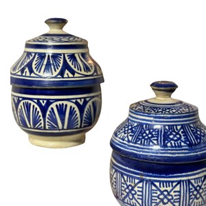 Puede incluir: Dos tarros de cerámica con tapas. Los tarros son azules y blancos con intrincados patrones geométricos. Las tapas tienen un pequeño pomo en la parte superior. Los tarros tienen forma redondeada y se asientan sobre una base de color crema.