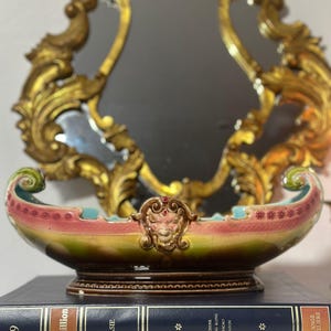 Könnte beinhalten: Eine dekorative, bootsförmige Keramikschale mit rosa, grünen und braunen Farbtönen. Die Schale hat ein detailliertes, verziertes Design und steht auf zwei dunkelblauen Büchern. Im Hintergrund befindet sich ein goldgerahmter Spiegel.