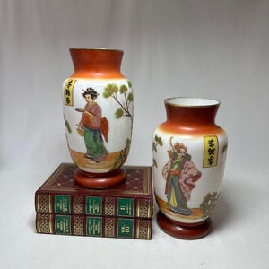Może przedstawiać: Dwa dekoracyjne wazony ceramiczne z białą podstawą, pomarańczową obwódką i drewnianą podstawą. Każdy wazon przedstawia malowaną postać azjatycką i drzewo. Wazony są wystawione na dwóch antycznych książkach z czerwonymi okładkami.