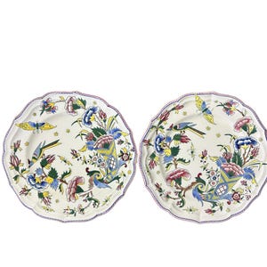 Puede incluir: Dos platos blancos con borde festoneado con un diseño floral y de pájaros. Los platos presentan un colorido patrón de flores y pájaros azules, rosas, amarillos y verdes. Los bordes están adornados con un borde morado.