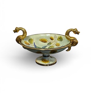 Può includere: Una ciotola decorativa in ceramica per centrotavola con esterno azzurro e design floreale. La ciotola ha manici dorati a forma di cigno e una base a piedistallo. L'interno presenta foglie e fiori dipinti di giallo, verde e blu.