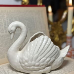 Puede incluir: Una figura de cisne de cerámica blanca con cuello curvo y diseño detallado de plumas. El cisne está sobre un libro abierto. Es un objeto decorativo.
