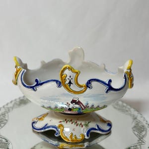 Puede incluir: Un cuenco de pedestal de cerámica blanca con borde festoneado, adornado con detalles azules y dorados. El cuenco presenta una escena pintada y se asienta sobre una base a juego. El cuenco se exhibe sobre una bandeja de espejo decorativa.