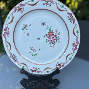May include: A white porcelain plate with a floral design. The plate features a central bouquet of flowers in shades of pink, orange, and blue. The rim is decorated with a repeating pattern of flowers and ribbons in pink, brown, and green.