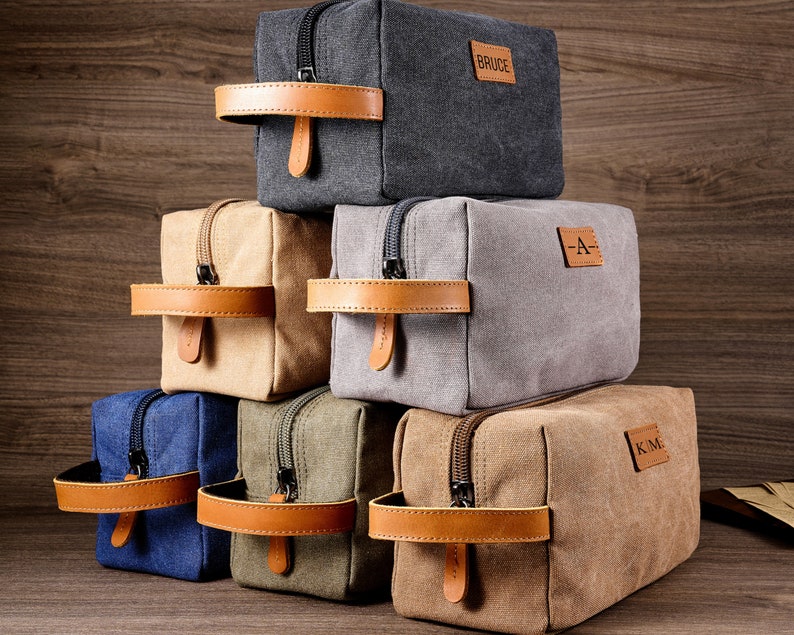 Puede incluir: Seis neceseres de lona con asas de cuero y cierres de cremallera. Las neceseres son de varios colores, incluyendo gris, beige, azul y verde. Cada neceser tiene un parche de cuero personalizado con iniciales o un nombre.