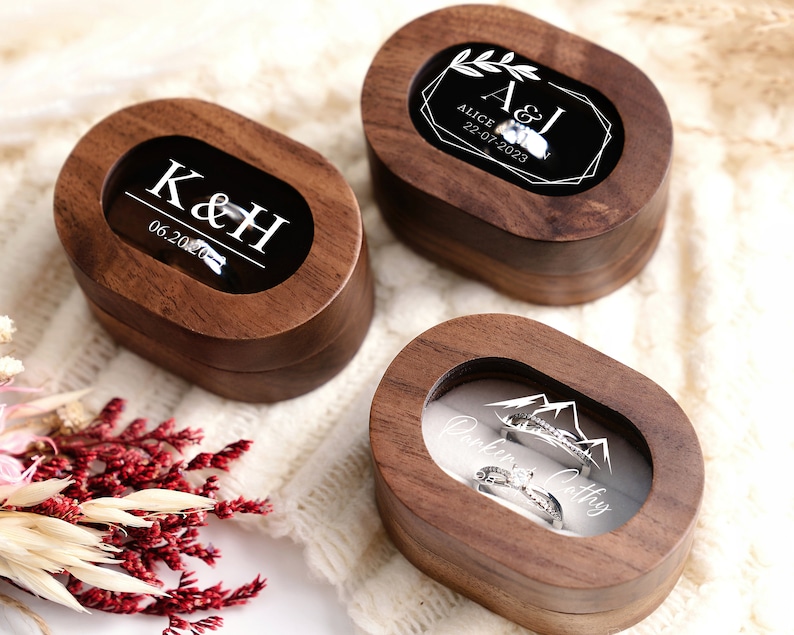 May include: Three wooden ring boxes with clear glass lids. The first box has the initials "K&H" and the date "06.20.20". The second box has the initials "A&I" and the date "22.07.2023". The third box has the words "Parker & Cathy" and a mountain range design.