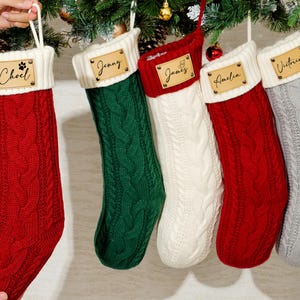 Peut inclure: Cinq bas de Noël en tricot torsadé rouges, verts, blancs et gris, chacun avec un revers blanc et une étiquette de nom en bois. Les bas sont suspendus à une branche de sapin.