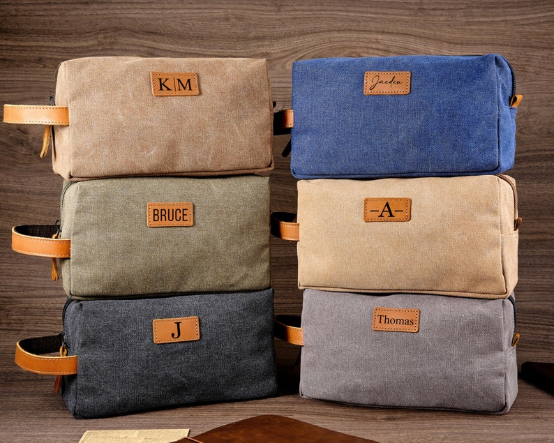 Puede incluir: Cinco bolsas de aseo de lona apiladas una encima de la otra. Las bolsas son de diferentes colores, incluyendo beige, azul, verde, gris y negro. Cada bolsa tiene un parche de cuero marr&oacute;n con un nombre o iniciales. Los nombres en las bolsas son "Kim", "Jaden", "Bruce", "A" y "Thomas".