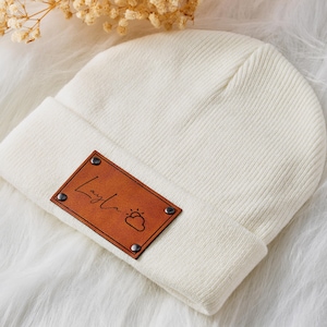Puede incluir: Gorro de punto blanco con un parche de cuero marrón que dice "Layla" con un gráfico de sol y nube.