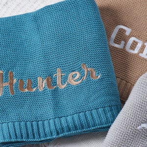 May include: A teal blue knitted baby blanket with the name "Hunter" embroidered in gold thread.