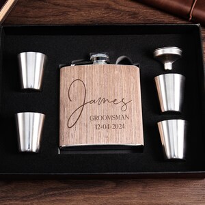 May include: A wooden box with a personalized stainless steel flask, a funnel, and four shot glasses. The flask is engraved with the name "James", the title "Groomsman", and the date "12-04-2024".