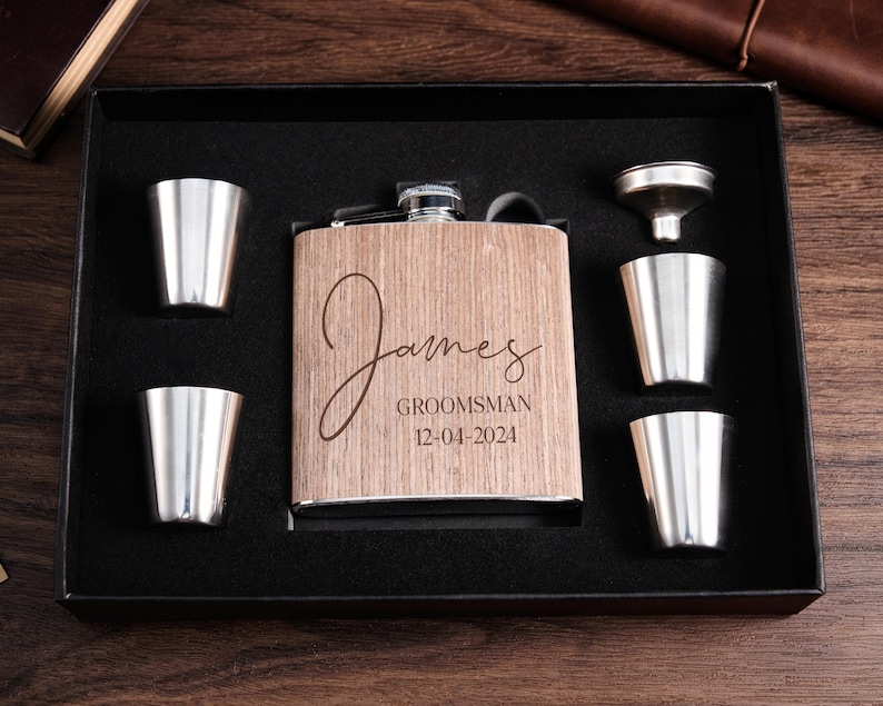 May include: A wooden box with a personalized stainless steel flask, a funnel, and four shot glasses. The flask is engraved with the name "James", the title "Groomsman", and the date "12-04-2024".