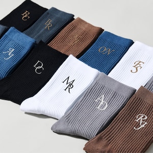 Può includere: Una collezione di calzini a coste in vari colori, tra cui nero, bianco, blu e marrone. Ogni calzino presenta iniziali ricamate in un colore a contrasto. I calzini sono disposti in un motivo diagonale, mostrando le diverse opzioni di colore.