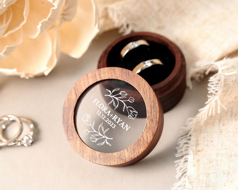 May include: A round wooden ring box with a clear lid and a floral design engraved on the inside. The lid has the text "FLORA + RYAN 12.12.2023" engraved on it.