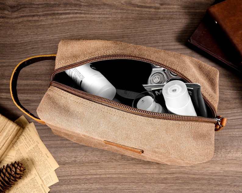 Puede incluir: Una bolsa de aseo de viaje de lona marr&oacute;n con cierre de cremallera y asa de cuero marr&oacute;n. La bolsa est&aacute; abierta y contiene varios art&iacute;culos de tocador, incluyendo un tubo de loci&oacute;n blanco y una maquinilla de afeitar plateada.