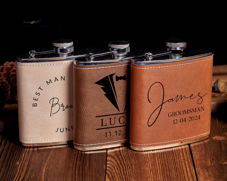 May include: Three personalized leather-wrapped stainless steel hip flasks. The flasks are brown, tan, and beige. The text on the flasks reads "Best Man", "Luc 11.12", and "James Groomsman 12-04-2024".