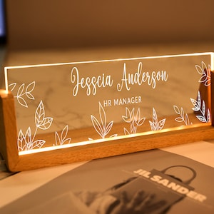 Könnte beinhalten: Ein hölzerner Schreibtisch-Namensschild mit einem transparenten Acryleinsatz. Der Einsatz ist mit dem Namen "Jesssica Anderson" und dem Titel "HR Manager" in Weiß graviert. Das Namensschild ist mit einem floralen Design verziert.