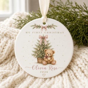 Pode incluir: Um enfeite redondo de cerâmica branca pendurado por uma fita creme. O design apresenta uma árvore de Natal, um urso de peluche a segurar uma estrela e o texto "MY FIRST CHRISTMAS", "Olivia Rose", "2026" e um pequeno coração impresso.