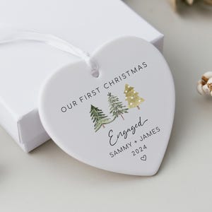 Könnte beinhalten: Ein weißer herzförmiger Weihnachtsschmuck mit dem Text "OUR FIRST CHRISTMAS" und "Engaged" mit Namen und dem Jahr 2024. Der Schmuck zeigt Weihnachtsbäume im Aquarellstil und ein weißes Band zum Aufhängen. Er liegt auf einer weißen Schachtel.