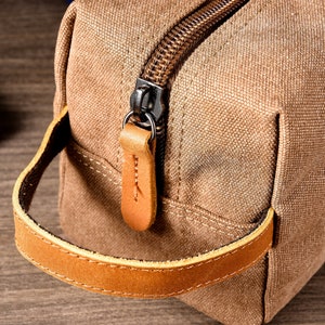 Puede incluir: Un neceser de lona marr&oacute;n con asa de cuero y cierre de cremallera.