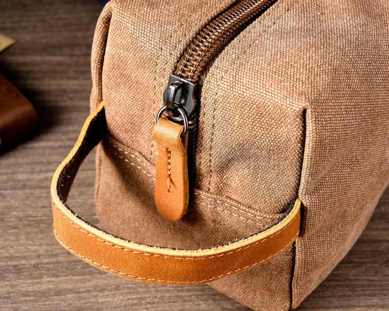 Puede incluir: Un neceser de lona marr&oacute;n con asa de cuero y cierre de cremallera.