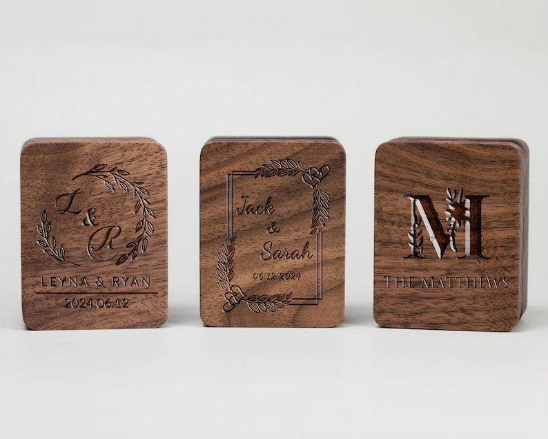 May include: Three wooden boxes with engraved designs. The first box has the initials "L & R" with a floral wreath and the text "Leyna & Ryan 2024.06.12". The second box has the text "Jack & Sarah 06.12.2024" inside a rectangular frame with floral accents. The third box has the initial "M" with a floral design and the text "The Matthews".