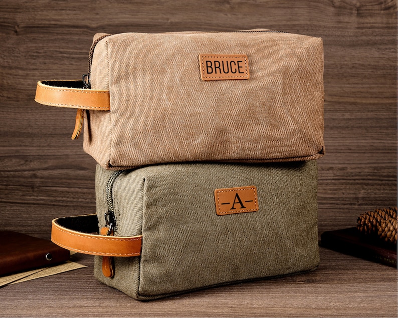 Puede incluir: Dos bolsas de lona con cremallera, asas de cuero y parches de cuero personalizados. La bolsa superior es marr&oacute;n con el nombre "BRUCE" en el parche. La bolsa inferior es verde con la letra "A" en el parche.