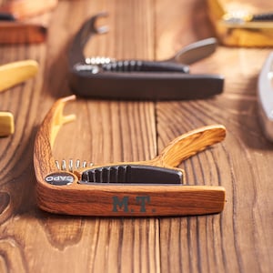 May include: A close-up of a wooden capo with the text "CAPO" and "M.T." engraved on it. The capo is on a wooden surface with other capos in the background.