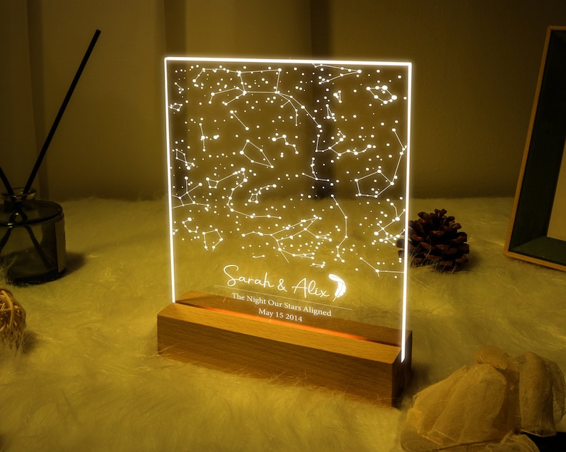 Puede incluir: Una luz nocturna personalizada con un mapa de estrellas en una base de madera. La luz muestra la constelaci&oacute;n de estrellas tal como aparec&iacute;an en una fecha espec&iacute;fica. El texto en la luz dice "Sarah & Alex, The Night Our Stars Aligned, May 15, 2014."