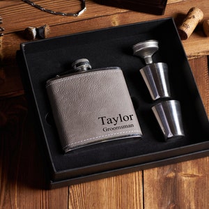 May include: A gray leather-bound flask with the name "Taylor" and the word "Groomsman" engraved on it. The flask is in a black gift box with two silver shot glasses and a funnel.