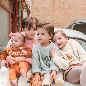 May include: Four young children in sweaters and pants pose together. One child wears an orange sweater with the word "tennis" embroidered on it. Another wears a light blue sweater with a green design. The other two children wear pink and cream sweaters.