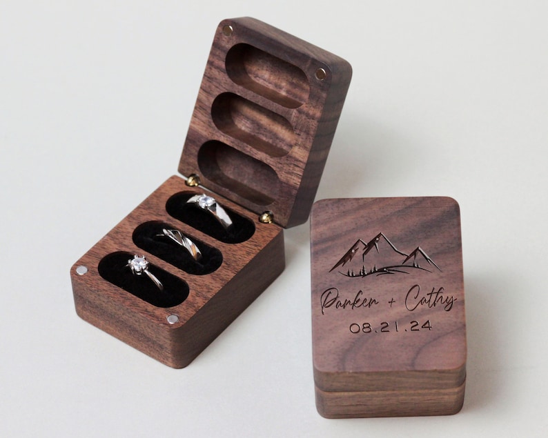 May include: A wooden ring box with a hinged lid. The box has four compartments for rings. The lid is engraved with a mountain range and the text "Parker + Cathy 08.21.24".