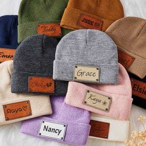 May include: A collection of knitted beanies with personalised leather patches. The beanies are in various colours, including grey, brown, pink, purple, green, and black. The patches feature names like "Teddy", "Jude", "Grace", "Kaya", "Elsie", "Raya", "Nancy", "Charlie", and "Emery".