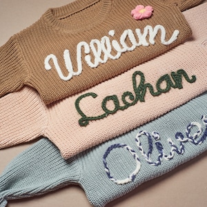 May include: Close-up of three knitted sweaters in brown, pink, and blue. Each sweater has a name embroidered on the front in a contrasting color: "William" in white, "Cachan" in green, and "Oliver" in blue and white. A pink flower is on the brown sweater.