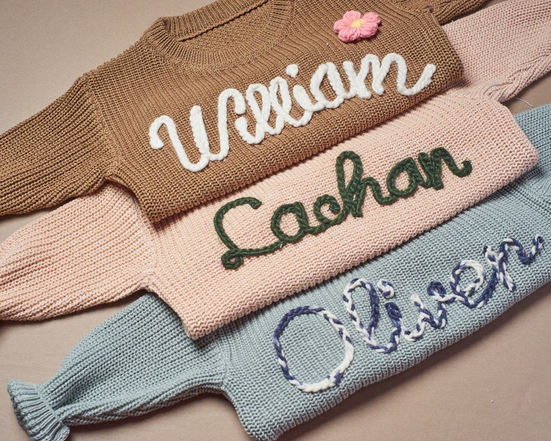 May include: Close-up of three knitted sweaters in brown, pink, and blue. Each sweater has a name embroidered on the front in a contrasting color: "William" in white, "Cachan" in green, and "Oliver" in blue and white. A pink flower is on the brown sweater.