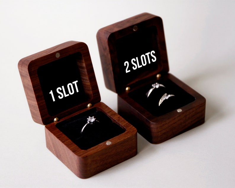 Op de afbeelding: Twee houten ringendoosjes met een zwart fluwelen interieur. Het kleinere doosje heeft &eacute;&eacute;n vakje en zegt "1 SLOT" op de deksel. Het grotere doosje heeft twee vakjes en zegt "2 SLOTS" op de deksel. Beide doosjes bevatten een ring.