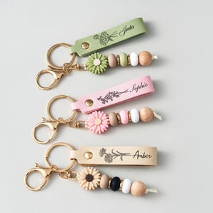 May include: Four keyrings with gold-coloured clasps and leather straps in green, pink, and tan. Each keyring features a floral design and a name: "Judes", "Sophie", and "Amber". The keyrings also have decorative beads and flower charms.