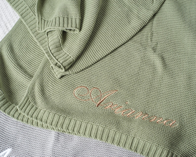 May include: A sage green knitted throw blanket with the word "Ariana" embroidered in gold thread.