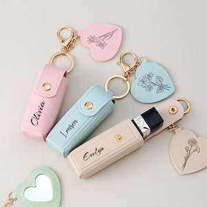 Könnte beinhalten: Drei rechteckige Lippenstiftetuis in Pastellrosa, Hellblau und Beige, jeweils mit goldenem Druckknopfverschluss und personalisiertem Namen. An jedem Etui befindet sich ein goldener Schlüsselring mit herzförmigem Anhänger und Blumenzeichnung.