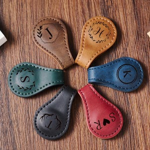 May include: Six leather keychains with different designs and initials. The keychains are brown, green, blue, black, and red. Each keychain has a different design, including a wreath, a heart, and antlers.