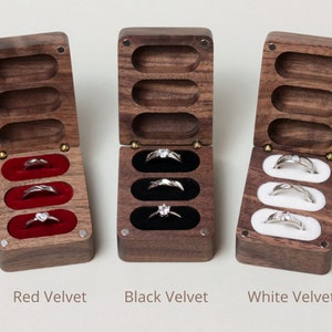 May include: Three wooden ring boxes with velvet interiors. The boxes are open and show three rings in each box. The boxes are lined with red, black, and white velvet.