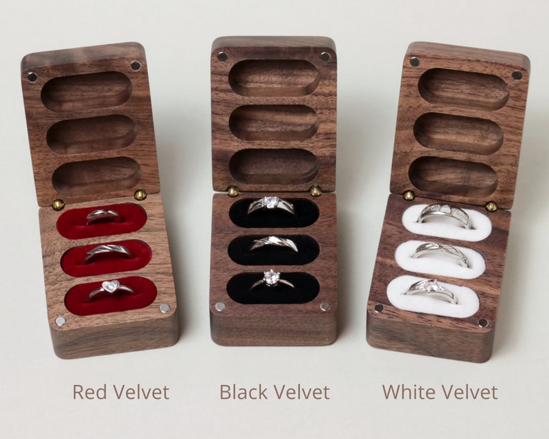 May include: Three wooden ring boxes with velvet interiors. The boxes are open and show three rings in each box. The boxes are lined with red, black, and white velvet.
