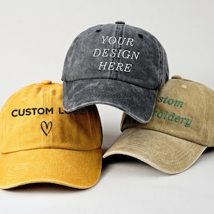 May include: Three baseball caps, one black with the text "YOUR DESIGN HERE", one yellow with the text "CUSTOM LOGO" and a heart, and one tan with the text "Custom Embroidery" in green.