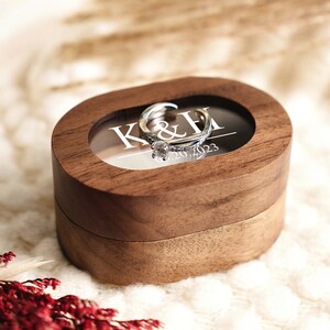 May include: A wooden ring box with a clear lid, showing a silver ring with a diamond. The box is engraved with the initials "K&H" and the date "07.20.2023".