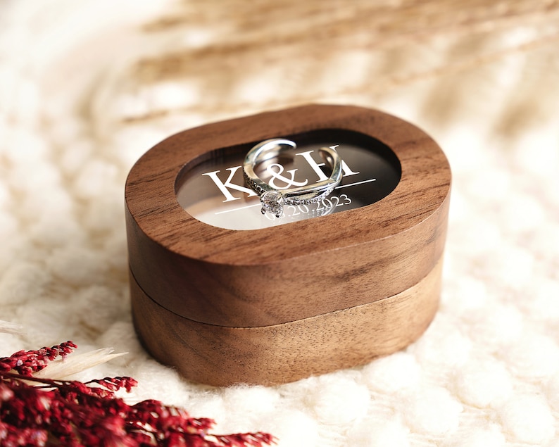 May include: A wooden ring box with a clear lid, showing a silver ring with a diamond. The box is engraved with the initials "K&H" and the date "07.20.2023".