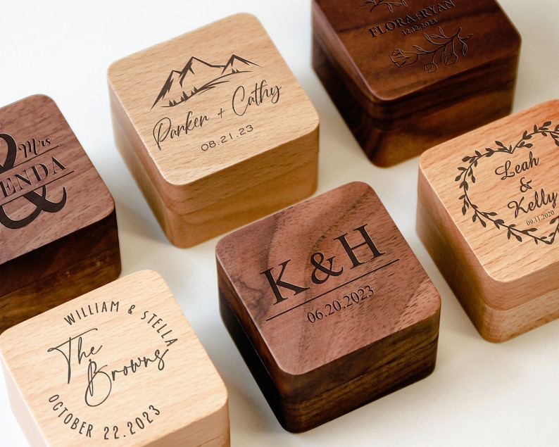 Op de afbeelding: Set van zes houten ringdozen met gegraveerde tekst en ontwerpen. De dozen zijn gemaakt van licht en donker hout en hebben een vierkante vorm. De tekst op de dozen bevat namen, datums en initialen.